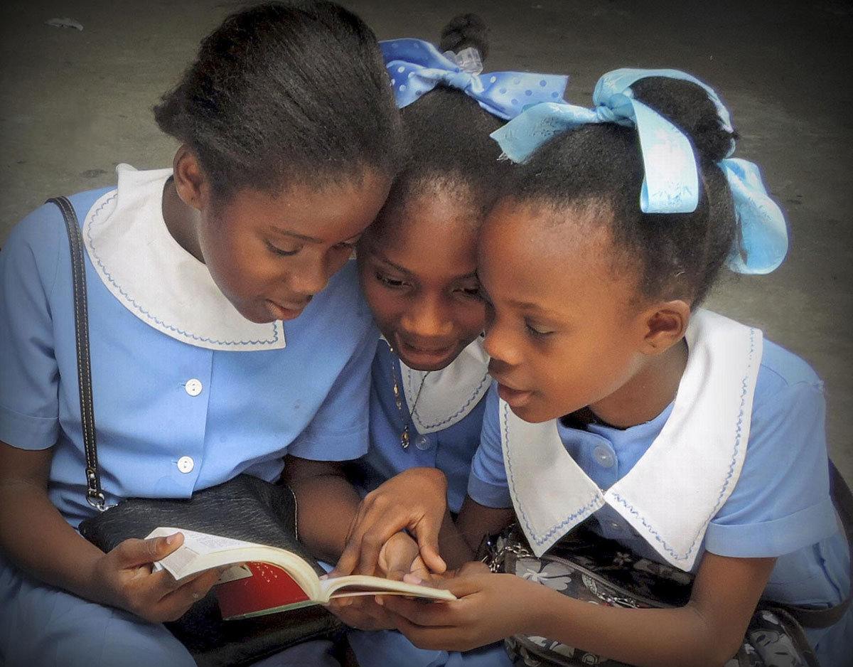 Three Haitian girls wearing light blue dresses with white collars read a book together.