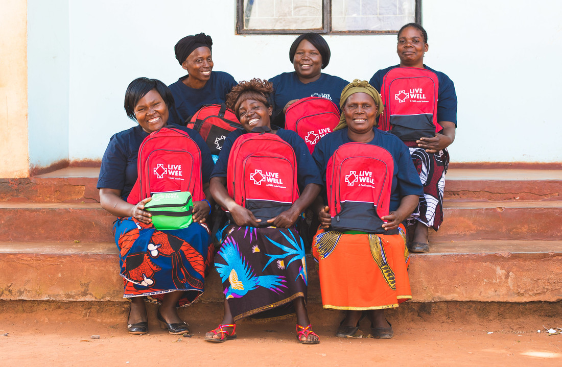 A group of six women sit on a set of stairs and smile with their red Live Well backpacks. Each woman is wearing a navy Live Well t-shirt and a brightly colored skirt.