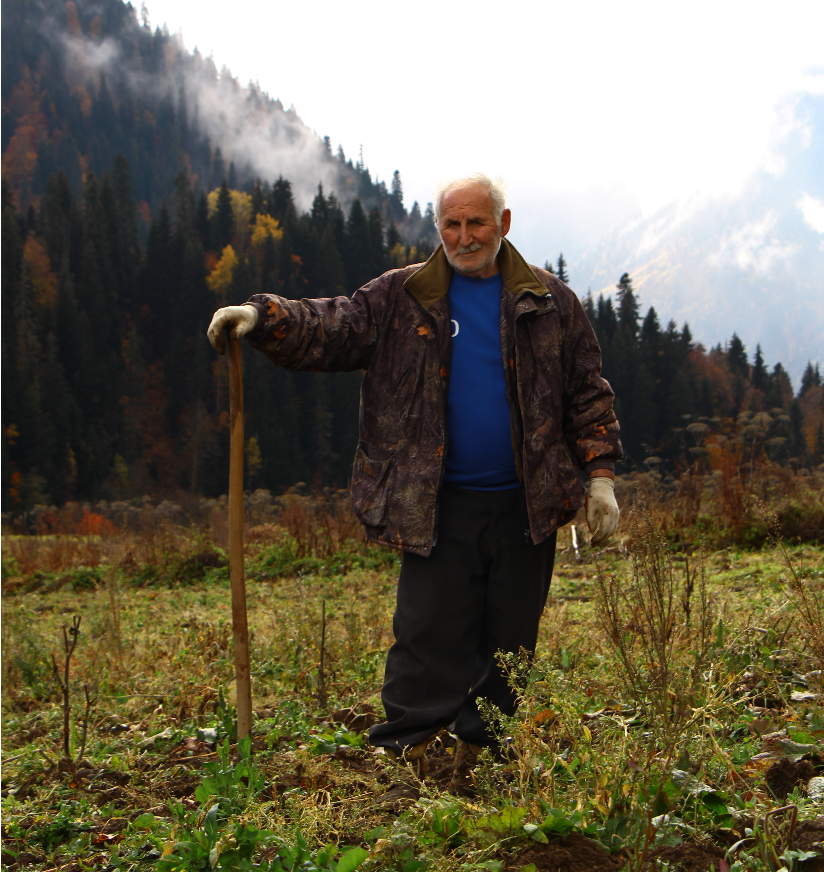 A middle-aged man stands in a field with his hand resting on a tall wooden staff. Behind him is a mountain covered with evergreens.